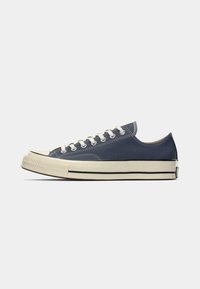 Adidași din pânză cu vârful jos, de culoare albastră închis, cu sireturi albe, capac de cauciuc pe vârf, talpă intermediară bej și o bandă de contrast neagră pe talpă.