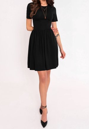 Zwarte jurk met korte mouwen en een aansluitend bovenlijfje, met een uitlopende rok, gemaakt van zacht materiaal. Gecombineerd met zwarte puntige hakken en gelaagde kettingen.