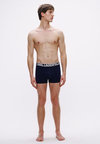 Granatowe bokserki z bawełny z miękkim paskiem w pasie, na którym znajduje się biały napis "KARL LAGERFELD". Model stoi boso, pokazany z profilu.