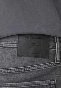 Grå jeans med en svart läderpatch som har präglad text, sydda sömmar och en lätt lyster i tygtexturen.