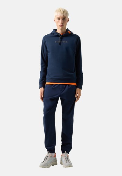 Mladý člověk s krátkými blond vlasy, oblečený v tmavě modré mikině s kapucí a teplácích, z nichž vykukuje oranžové tričko, a světlých šedých teniskách.