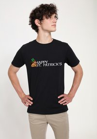Černé bavlněné tričko s nápisem "ŠŤASTNÝ PATRICKŮV DEN" v bílém písmu a grafikou skřítka s zeleným kloboukem.