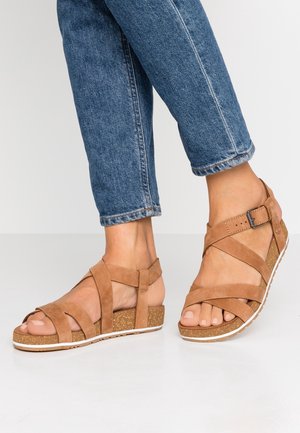 Voeten met beige suède sleehakken met gekruiste bandjes en een gesp, gecombineerd met blauwe denim jeans die boven de enkels zijn afgeknipt.