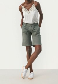 Débardeur blanc à bordure en dentelle associé à un short kaki et des baskets blanches. Le short possède des poches latérales et une coupe droite.