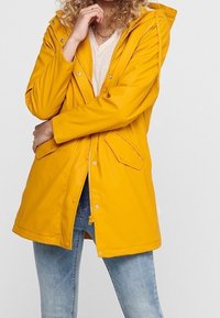 Imperméable jaune avec capuche, fermeture éclair frontale, poches à boutons-pression, et design longueur genou, assorti à un jean bleu clair.