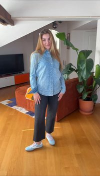 Grūtniece ar gariem blondiem matiem stāv viesistabā, valkājot zilu rakstainu kreklu, melnas bikses un gaiši zilas sporta kurpes.