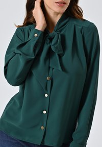 Blusa teal realizzata in tessuto liscio, con design a collo legato, chiusura con bottoni e dettagli in metallo dorato. Maniche lunghe con polsini aderenti.