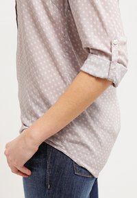 Camicia grigia chiaro con motivo floreale, polsini abbottonati e orlo leggermente curvato. Realizzata in tessuto morbido con una texture liscia.