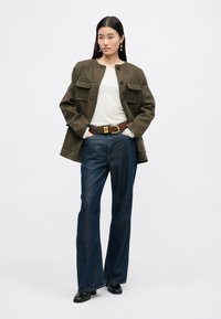 Brązowa kurtka w kratę z dwiema przednimi kieszeniami, biały top, ciemnoniebieskie dzwony z denimu i brązowy pasek ze złotym okuciem. Czarne botki sięgające kostki.