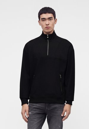 Ung man som bär en svart tröja med halvlång dragkedja i kragen och dragkedjeförsedda framfickor, ihop med blekta grå jeans, mot en enfärgad bakgrund.
