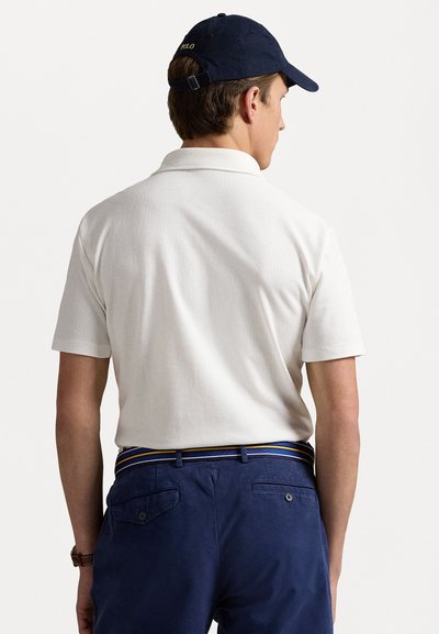 Hvid kortærmet polo-shirt, marineblå kasket, blå bukser med et mønstret bælte. Fokus på rygvisning, der fremhæver udskæringen og stofstrukturen.