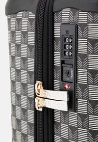 Valise rigide avec un motif géométrique gris, dotée d'un cadenas, de tirettes dorées et d'une surface texturée pour une meilleure prise.