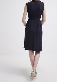 Marinblå ärmlös klänning med figursydd bodice och A-linjeformad kjol, med sidofickor och en mjuk textur. Matchad med metalliska klackar.