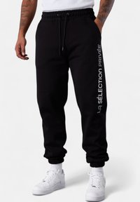 Man wearing black joggers with "LA SÉLECTION PRIVÉE" text on the right leg, white socks, and white sneakers, standing with hands in pockets.