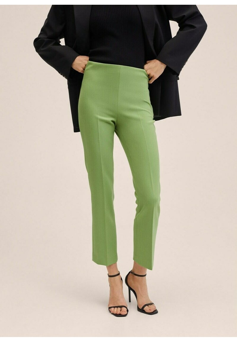 Femme portant un pantalon vert citron sur mesure, un haut noir côtelé, un blazer noir et des sandales à talons aiguilles noires, les mains ajustant les côtés de sa veste.