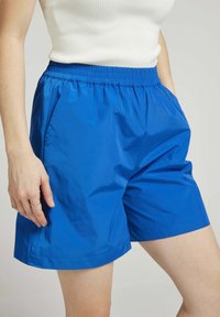 Shorts en nylon bleu avec une taille élastique et des poches latérales. Texture lisse et design simple, atteignant juste au-dessus du genou.
