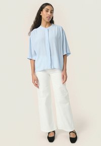 Blusa azzurra chiara a maniche corte con chiusura a bottoni, caratterizzata da una vestibilità comoda e da una texture morbida. Abbinata a pantaloni bianchi a palazzo e scarpe nere.