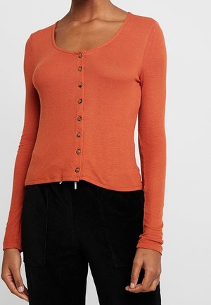 Femme portant un haut orange ajusté à manches longues boutonné, associé à un pantalon noir, vue du cou aux hanches sur un fond uni.