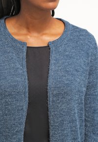 Cardigan blu lavorato a maglia con scollatura rotonda. Presenta una finitura morbida e testurizzata e un design aperto sul davanti. Indossato sopra una maglia nera.