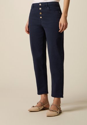 Pantaloni blu navy vita alta con quattro bottoni metallici indossati con ballerine beige a punta con cinturini intrecciati.