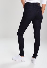 Jeans skinny de mezclilla oscura con una cintura alta, que presentan detalles de costura sutiles y un diseño de bolsillo trasero. Combinados con zapatillas deportivas blancas.