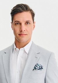 Mężczyzna z krótkimi ciemnymi włosami, ubrany w jasnoszary żakiet, białą koszulę i wzorzysty niebieski poszetkę, patrzący bezpośrednio w kamerę.