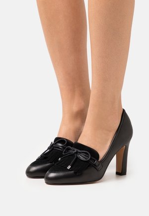 Black leather heeled shoes with tassel detailing and a bow on the front; smooth texture with a pointed toe and mid-height heel.