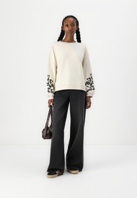 Overdimensjonert kremfarget sweatshirt med svarte intrikate mønstre på ermene, kombinert med vide svarte jeans og svarte sneakers med lyse såler.