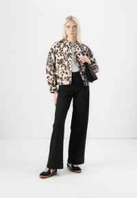 Chaqueta de estampado animal en beige y marrón, blusa blanca, pantalones negros de talle alto y zapatillas negras con detalles en blanco, sosteniendo un bolso negro.