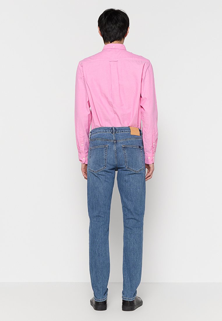 Camisa de manga larga rosa con cuello abotonado, combinada con jeans de mezclilla azules que tienen una etiqueta de cuero en la parte trasera de la cintura. Zapatos negros.