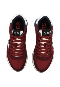 Un paio di sneakers rosse e bianche con lacci bordeaux e linguette nere che presentano simboli geometrici bianchi, viste dall'alto.