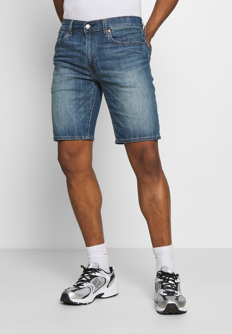 Denim shorts dalam warna biru sedang, dengan potongan santai dan pinggiran yang dijahit. Dipadukan dengan sepatu lari hitam dan kaus kaki putih.