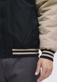 Black and beige bomber jacket with a smooth finish, featuring snap buttons, striped ribbed cuffs, and a contrasting waistband.