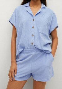 Žena v světle modré košili s krátkým rukávem a kapsou, která má zapínání na knoflíky, a odpovídajícími šortkami na gumu, stojící s jednou rukou v kapse.