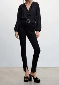 Blouse noire à col en V avec détail noué, associée à un pantalon noir ajusté avec des fentes lat érales. Complété par des sandales plateformes noires à talons hauts.