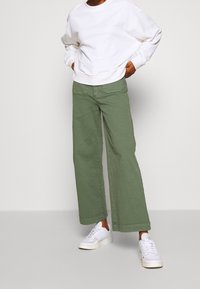 Witte sweatshirt en wijde groene broek, gecombineerd met witte sneakers. De kledingstukken hebben een ontspannen pasvorm en eenvoudige, strakke lijnen.