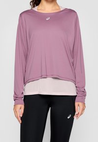 Top atletico leggero a maniche lunghe in color malva con orlo arrotondato. Presenta un design a strati e un piccolo logo sul petto. Accoppiato con leggings neri.