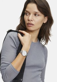 Silberne Metalluhr mit einem runden weißen Zifferblatt, einfachen Stundenmarkierungen, am Handgelenk getragen; Person in einem gerippten grauen Langarmoberteil.