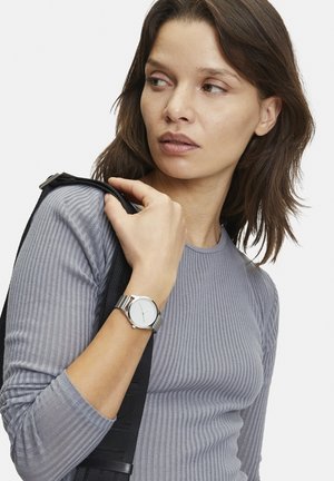 Montre en métal argenté avec un cadran blanc rond, marqueurs horaires simples, portée au poignet ; personne portant un haut à manches longues gris à côtes.