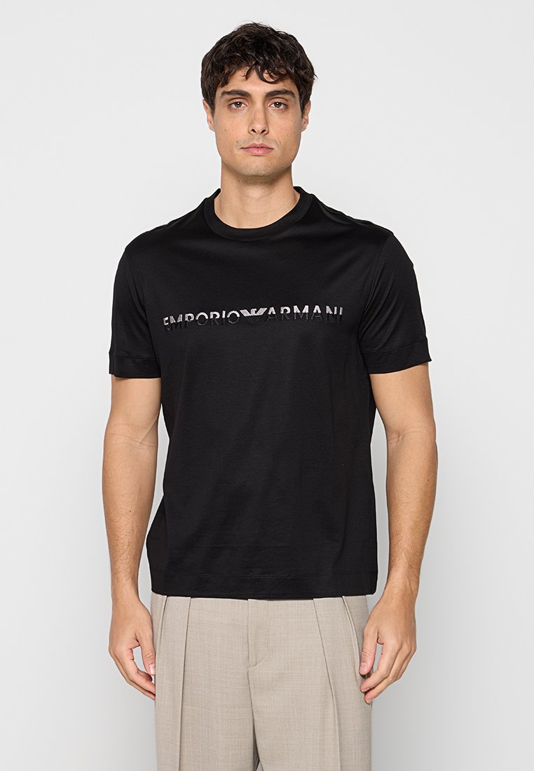 Hombre con camiseta negra de Emporio Armani y pantalones plisados beige, de pie frente a un fondo liso gris claro.
