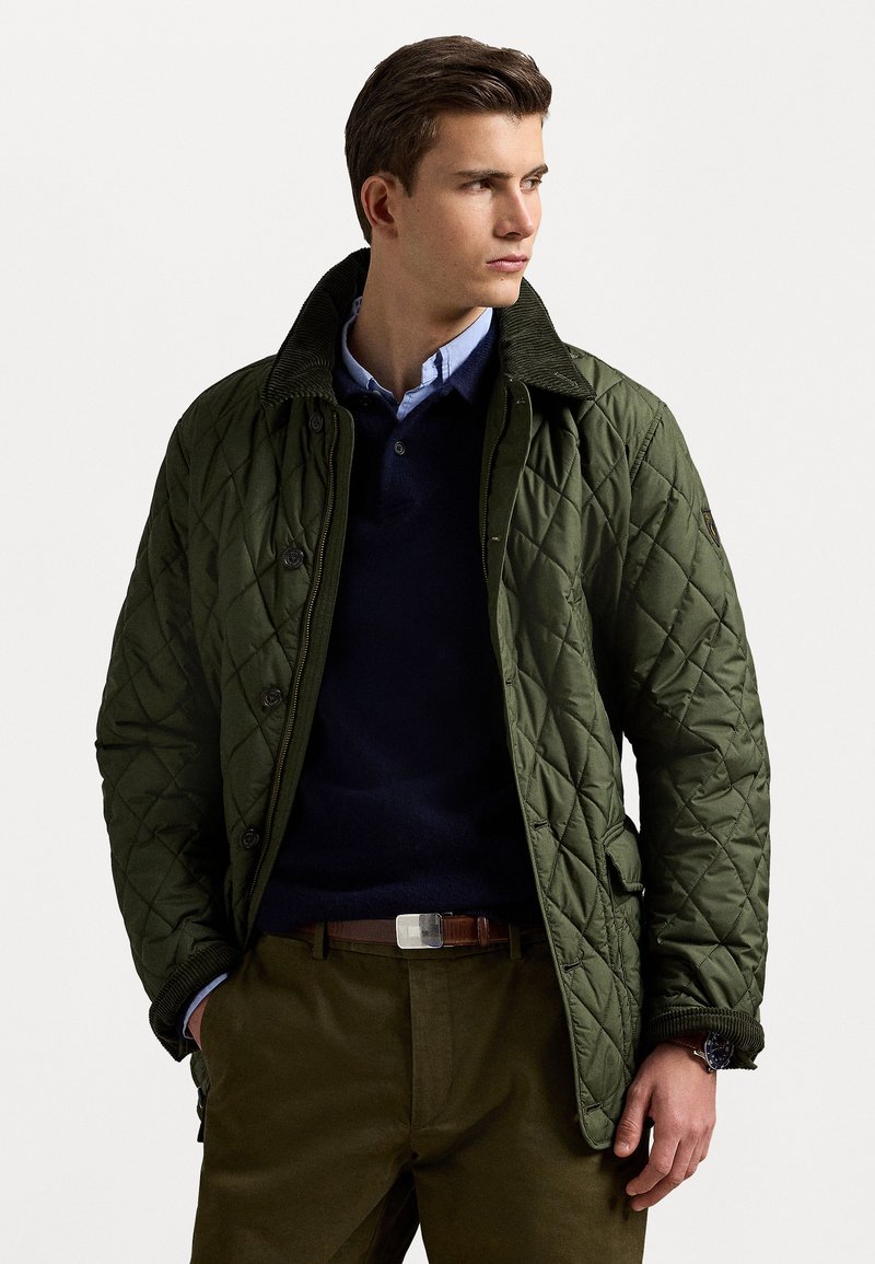 Khaki green quilted jacket with a corduroy collar, two front pockets, and button closure, worn over a navy sweater and light blue shirt.