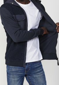 Marinblå zip-up jacka med en texturerad, fleece-liknande framdel. Inkluderar en vit t-shirt under och blå jeans. Ozip-ad för att visa inredningsdetaljer.