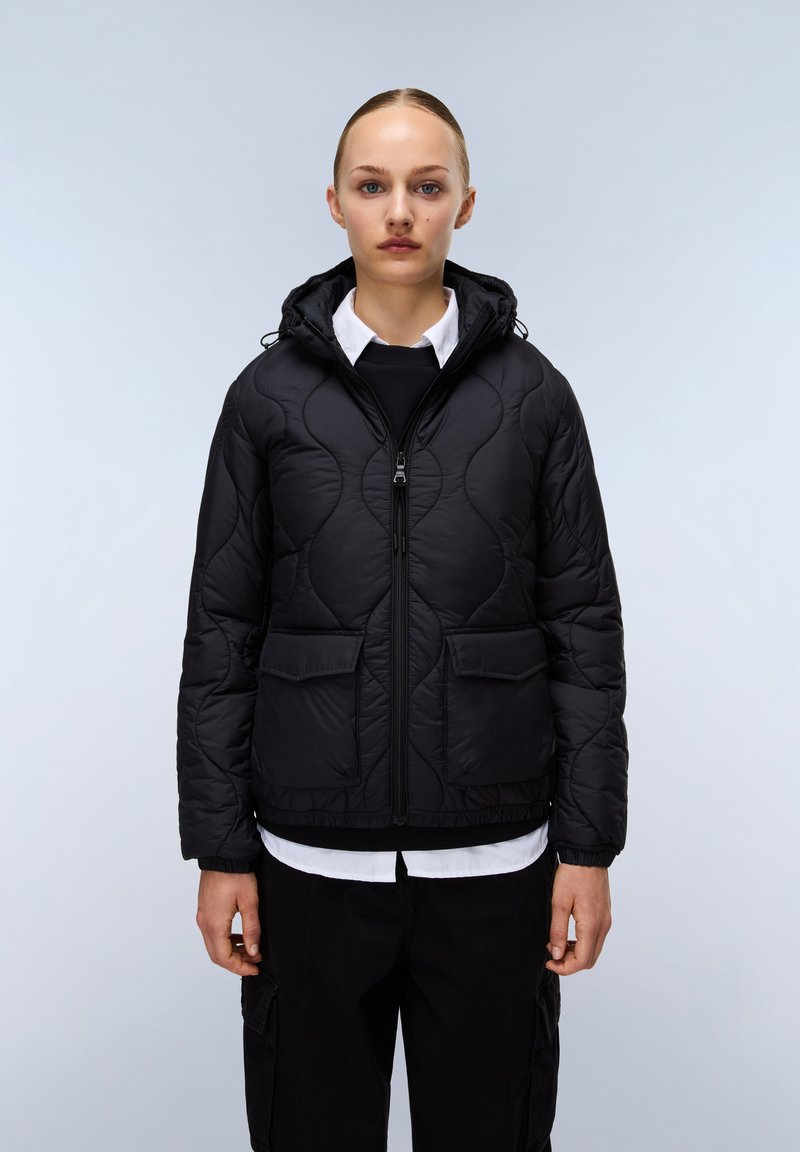 Svart quiltad puffjacka med huva, frontdragkedja och två stora fickor. Har en slät yta och elastisk nederkant, lager på lager över en vit skjorta.