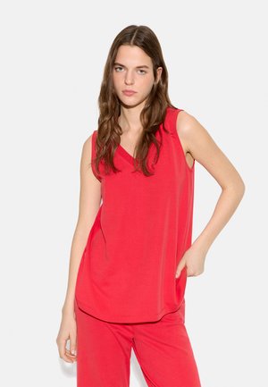 Kobieta o długich brązowych włosach, ubrana w czerwony, bezrękawnikowy top z dekoltem w serek i pasujące czerwone spodnie, stojąca z jedną ręką w kieszeni.