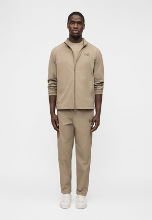 Egy férfi áll, bézs színű, zipzáras kabátból, nadrágból álló, az EA7 márkájú melegítőben és fehér sportcipőben, egyszínű fehér háttér előtt.