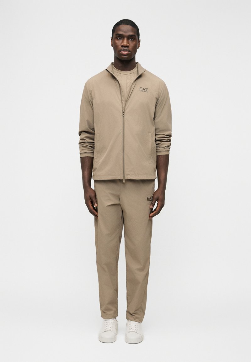 Άνδρας στέκεται φορώντας μπεζ ασορτί φόρμα EA7 με τζάκετ με φερμουάρ, παντελόνια και λευκά αθλητικά παπούτσια, μπροστά σε μονόχρωμο λευκό φόντο.