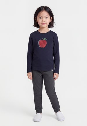 Jeune fille debout portant un haut bleu marine à manches longues avec un motif de pomme rouge, un pantalon gris et des chaussures blanches à enfiler sur un fond uni.