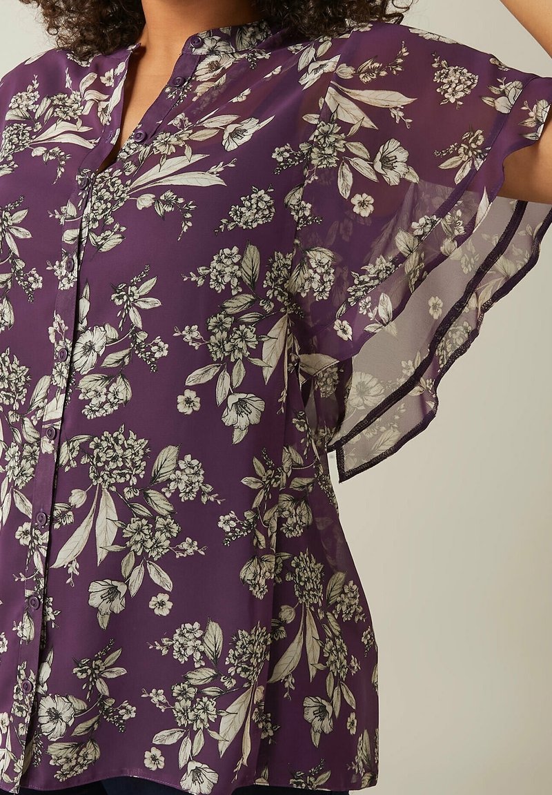 Blouse violette transparente avec imprimé floral blanc, boutonnée sur le devant, manches volantes, portée par une personne avec le bras levé.