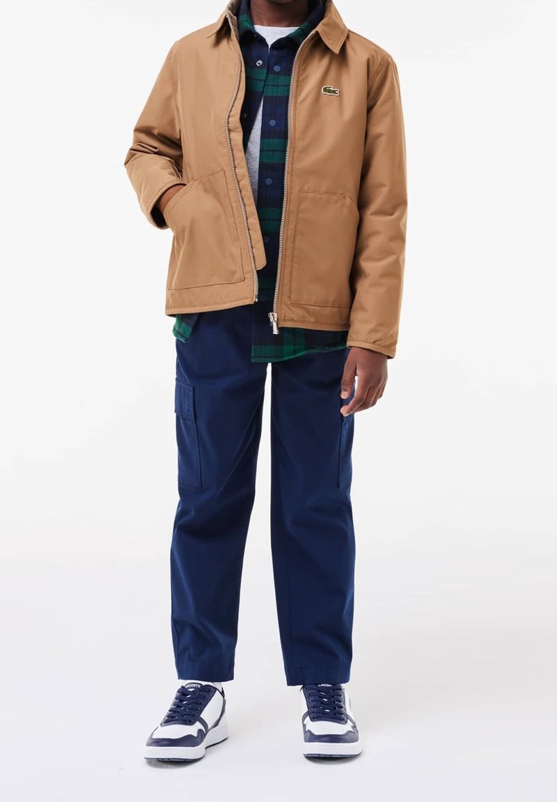 Homme portant une veste zippée couleur beige, une chemise à carreaux verte et bleu marine, un pantalon cargo bleu marine et des baskets blanches et bleu marine, debout sur fond blanc.