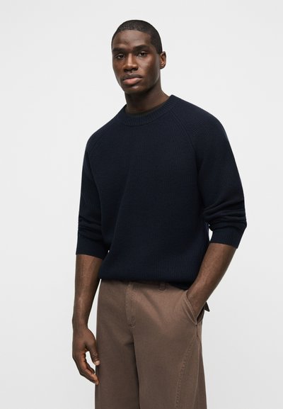 Άνδρας που φοράει μπλε σκούρο πλεκτό πουλόβερ με μακριά μανίκια και καφέ παντελόνι, στέκεται με το ένα χέρι στην τσέπη, μπροστά σε απλό φόντο.